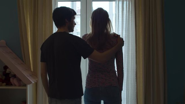 Silhouette of a resentful woman near the window. Her husband approaches her and puts hand on her shoulder in search of reconciliation. Wife angrily pushes his hand away