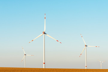 4 white wind turbines with a red stripe stand on a freshly plowed field. The rotors stands still.
