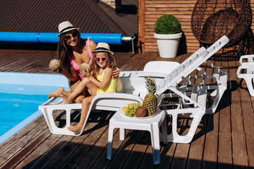 Mom with her daughter in hats and glasses posing on a sunbed holding a coconut in her hand.