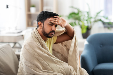 health, cold and people concept - sick young indian man in blanket having headache or fever at home - obrazy, fototapety, plakaty