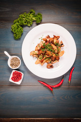 A plate of appetizing potato with mushrooms on a wooden table, among seasonings. Studio photography of food in the cooking industry, dark background.