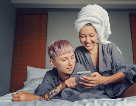 Two Beautiful Young Girlfriends Charming Lesbian Couple In Bathrobes Look At The Screen Of A Smartphone And Have Fun In The Hotel Room