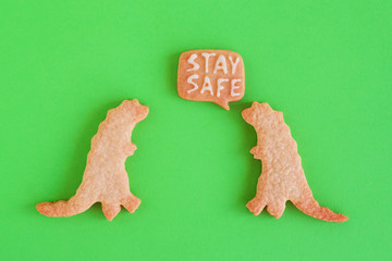 Two homemade cookies in shapes of dinosaurs with inscription ‘Stay safe’ on green background, top view. Sweet shortbread with white glaze. Social distancing concept.