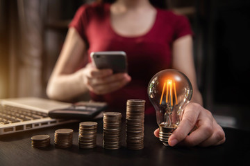 Business woman hand holding lightbulb with coins stack on desk. concept saving energy and money at office.