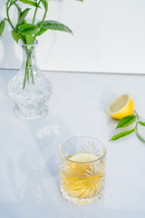 Crystal glass with yellow cocktail, vase with fresh greenery and half of lemon on marble background. Glass goblet with green tea, alcohol drink. Direct sunlight and shadows. Minimalism. Copy space.