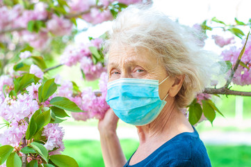 Ederly woman in a protective respiratory mask in a blossoming pink sakura tree. Quarantine, health,...