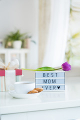 Mother's day background. Morning suprise - cup of tea with cookies, gift box, lightbox with words Best mom ever and tulip flower on the marble table with light interior view. Vertical card. Copy space