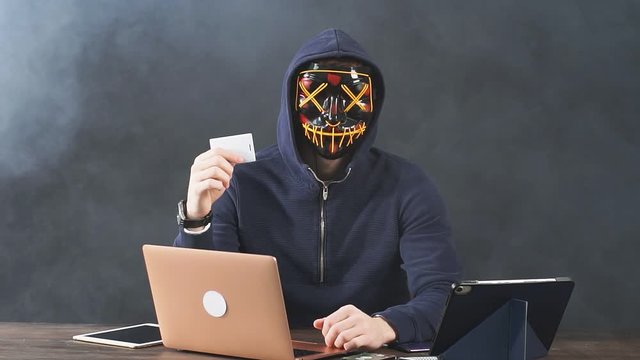 Portrait of a masked male hacker with a laptop and credit card in hand, looking at camera and giving thumbs up.