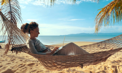 Young woman working on laptop lying in hammock at sand beach of tropical island. Freelance outdoor...