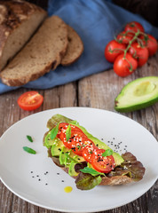 Fototapeta na wymiar Toast with freshly baked organic bread, olive oil, lettuce, avocado, sesame seeds and cherry tomatoes on a white plate on a dark wooden background. Close-up, lunch concept