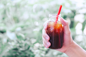Close up hand holding bottle glass of americano coffee with green nature background, vintage tone