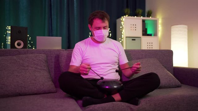 a man in a protective mask sits at home and plays on the hapi drum