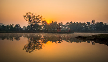 Colourful and warm glow of sunlight reflected on a serene lake during sunrise. A calm and spiritual scene with a temple and trees at Tal Pokhara, Rampur, Palpa, Nepal. 