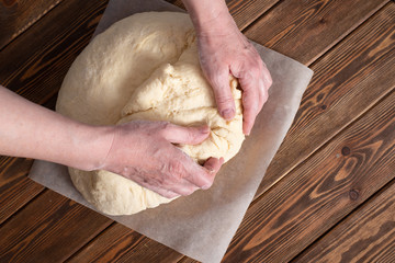 A piece of dough on parchment lies on a wooden table. person kneads the dough, part of the recipe