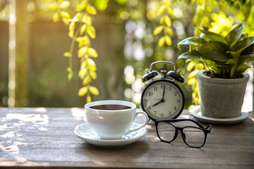 Hot Coffee​ in​ white​ cup​ with​ eye glass​ and alarm clock on​ wooden​ table​ and​ blurred​ background​ at​ morning​ time.