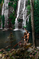 Fototapeta na wymiar Beautiful couple at a waterfall in Indonesia. A couple in love travels around the island of Bali. A man and a woman kiss at the waterfall. Travel to Asia. Honeymoon trip. Tourists at the waterfall