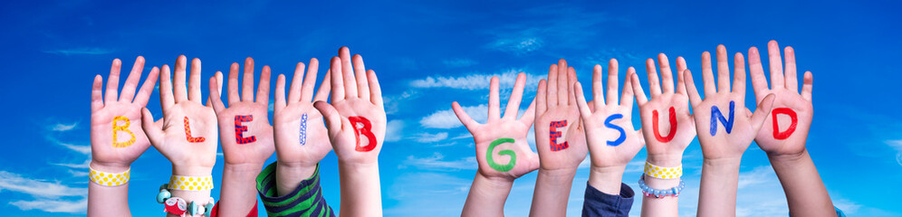 Kids Hands Holding Colorful German Word Bleib Gesund Means Stay Healthy. Blue Sky As Background