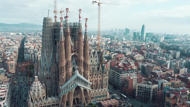 Aerial; drone view of main Barcelona attraction; majestic building towering over the rooftops of Eixample; sharp domes and unusual forms of great unfinished work of neo-gothic art; camera moves around