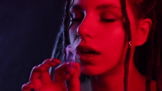 the girl exhales a pair of cannabis, smokes a cigarette with marijuana close-up, enjoys smoking. blows smoke in neon lights, dreadlocks and tattoos