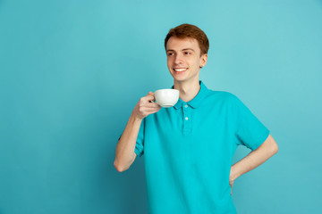 Enjoying coffee, delighted. Caucasian young man's portrait isolated on blue studio background, monochrome. Beautiful male model. Concept of human emotions, facial expression, sales, ad, trendy.