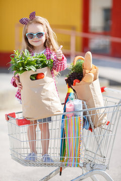 Adorable baby kid with trolley choosing fresh vegetables in local store. Girl with a big grocery cart in a parking lot near a store. A little girl with a big trolley buys fresh food and vegetables in 