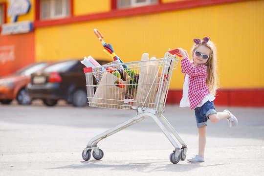 Adorable Baby Kid With Trolley Choosing Fresh Vegetables In Local Store. Girl With A Big Grocery Cart In A Parking Lot Near A Store. A Little Girl With A Big Trolley Buys Fresh Food And Vegetables In 