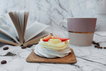 Homemade beautiful Breakfast. White dessert of meringue, cream and fresh strawberries, hot drink mug, open book and coffee beans on white marble surface