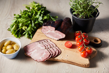 tasty ham on cutting board with greens, cherry tomatoes, olives on wooden table