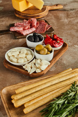 High angle view of boards with breadsticks, salami, rosemary, cheese and bowls with antipasto ingredients on brown