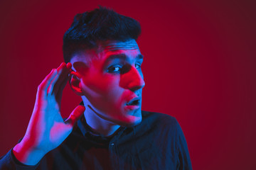 Listening to secrets with hand on ear. Caucasian young man's portrait isolated on red studio background in neon light. Concept of human emotions, facial expression, sales, ad, youth culture.
