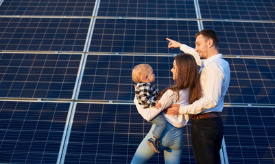 Back view of a beautiful young family, standing together holding a baby near photovoltaic solar...