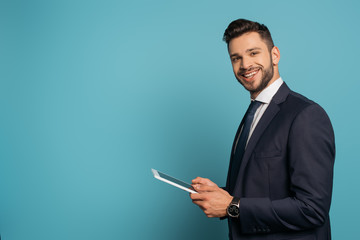 handsome businessman using digital tablet while smiling at camera on blue background