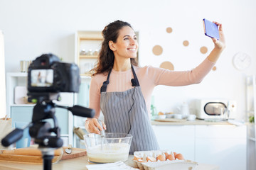 Attractive young adult woman wearing apron taking selfie while shooting video for her food vlog