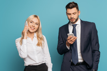 smiling businesswoman talking on smartphone near serious businessman chatting on blue background