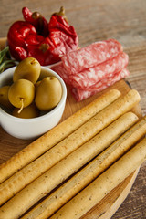 High angle view of breadsticks and salami slices near bowls with marinated chili peppers and olives on board on wooden background