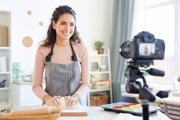 Horizontal medium portrait of cheerful young woman wearing apron kneading dough on camera for her food blog