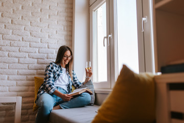A beautiful caucasian young woman with a glass of wine by the window reading a book