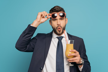 surprised businessman touching sunglasses while holding cocktail glass with orange juice isolated on blue
