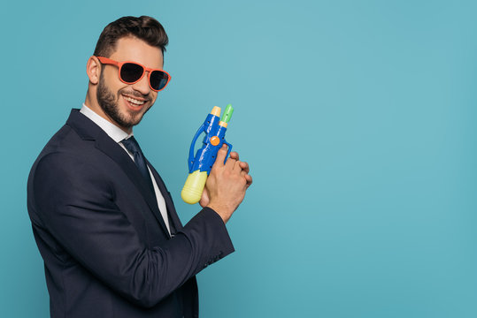 joyful businessman holding water gun while looking at camera isolated on blue