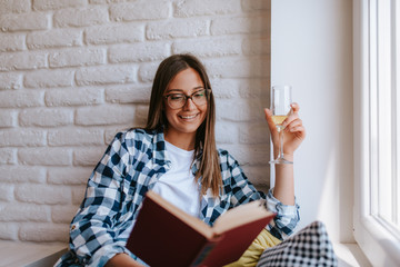 A beautiful caucasian young woman with a glass of wine by the window reading a book