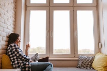 A beautiful caucasian young woman with a glass of wine by the window reading a book