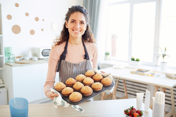Modern Caucasian woman holding mold with hot cupcakes taken out of baking oven looking at camera...