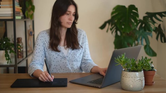 Professional young graphic designer sitting at her desk uses laptop at home. She retouches videos and photos in an image editing software using drawing tablet on a warm day in a room full of plants