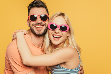 happy handsome man and attractive blonde woman in sunglasses embracing isolated on yellow