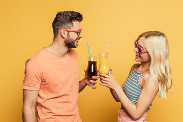 smiling young couple in sunglasses clinking cocktail glasses on yellow background
