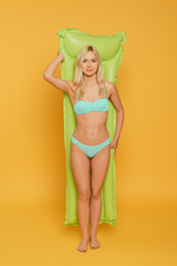 full length view of attractive, blonde girl in swimsuit holding inflatable mattress on yellow background