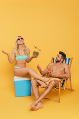 excited blonde girl sitting on portable fridge near handsome man holding slice of watermelon in deckchair on yellow background