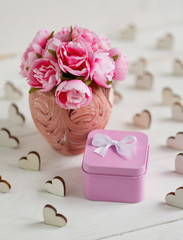 Beautiful composition with small square metallic pink gift box and delicate roses in pink vase on white wooden background
