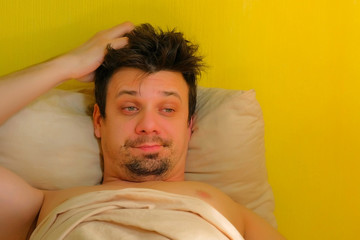Fototapeta na wymiar Sleepy tired bearded man with hangover wakes up in bed looking at camera, portrait closeup. Young man with headache in the morning after waking up. Tired sick guy. Yellow wallpaper background.