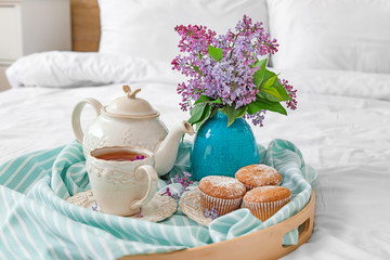 Vase with beautiful lilac flowers, tea and muffins on tray in bedroom